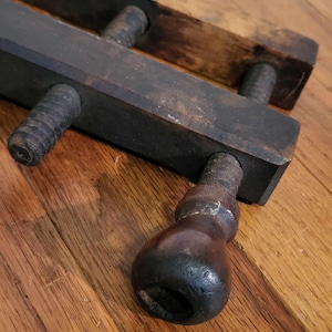 May include: A vintage wooden clamp with a dark brown finish. The clamp features two parallel rectangular bars connected by threaded rods and a handle. The wood shows signs of wear, suggesting age and use. The tool is designed for woodworking or similar crafts.