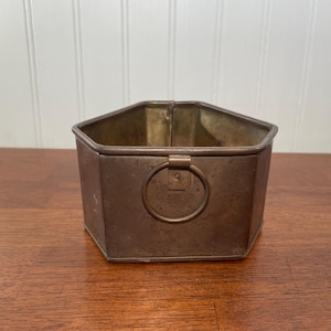 May include: A hexagonal metal container with a weathered bronze-toned finish. The vessel features a flat base, straight vertical sides, and a single circular metal ring handle attached to the center of the front panel with a small square plate.
