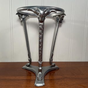 May include: A silver-toned metal stand featuring three legs, each topped with a sculpted ram head. The legs are connected by a circular ring at the top and a triangular base at the bottom. The legs have a textured, floral-like pattern along them.