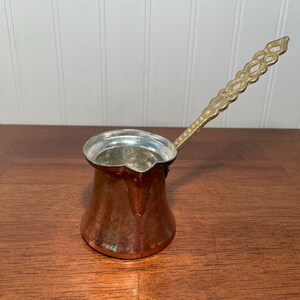 May include: A copper coffee pot with a wide, flared base and a narrow neck featuring a silver-toned interior lining. The pot includes a long, decorative brass handle with an intricate, repeating openwork pattern extending from the side rim.