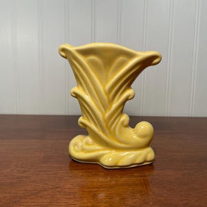 May include: A yellow ceramic vase with a stylized, organic design. The vase has a flared top and a base with curved, flowing shapes. The vase is approximately 7 inches tall and is a solid, pale yellow color.