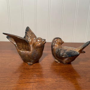 May include: Two ceramic bird figurines with a glossy brown and black mottled glaze. One bird is depicted with its wings spread upward, while the other bird is shown in a resting position with its wings folded against its body and head turned.