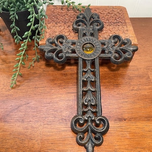 May include: A dark metal wall cross featuring intricate scrollwork and fleur-de-lis accents. The center of the cross is adorned with a single, circular, amber-colored glass cabochon, providing a contrast to the textured, dark iron-like finish.