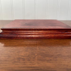 May include: A rectangular, stained wooden display base with a tiered, decorative edge. The wood has a rich, reddish-brown color and a smooth, polished finish. The base is designed to elevate and showcase items.