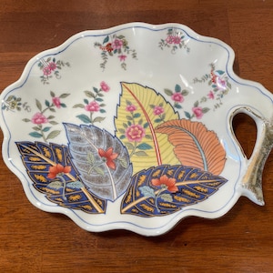 May include: A white ceramic leaf-shaped dish with a scalloped edge and a gold handle. The dish is decorated with colorful leaves in yellow, orange, and blue, and pink floral accents. The dish is approximately 10 inches long.