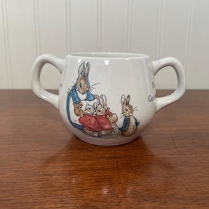 May include: White ceramic cup with two handles, featuring a colorful illustration of rabbits in a storybook style. The cup has a rounded shape and is likely designed for a child. The artwork includes rabbits in blue, red, and brown clothing.