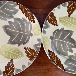 May include: Two white ceramic plates with a leaf design. The leaves are in shades of gray, brown, and light green. The plates are on a dark brown wooden surface. The plates are likely for serving food.