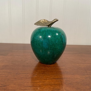 May include: A decorative apple figurine with a vibrant green body and a brass leaf stem. The apple is set on a wooden surface, showcasing its polished, glossy finish. The figurine is a unique home decor item.