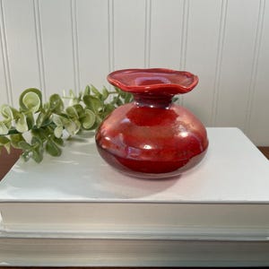 May include: A small, round, red ceramic vase with a flared top, sitting on a stack of white books. The vase has a glossy finish and a unique shape. A sprig of green leaves is placed near the vase.