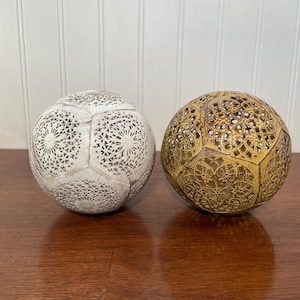 May include: Two decorative spheres with intricate, openwork designs. One sphere is white, the other is gold. The spheres are placed on a dark wooden surface, with a white paneled wall in the background. The spheres have a soccer ball shape.