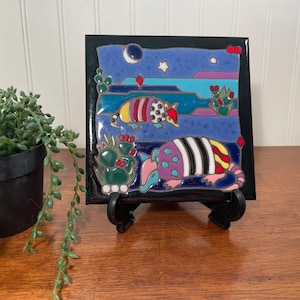 May include: A square ceramic tile featuring a colorful desert scene with two armadillos, cacti, a crescent moon, and stars against a blue background. The tile is framed in black and rests on a black plastic display stand on a wooden surface.