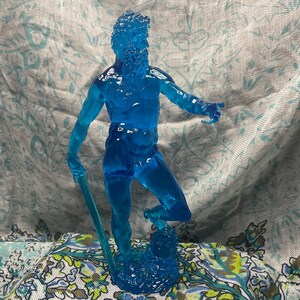 K&ouml;nnte beinhalten: Eine durchscheinende blaue Figur eines b&auml;rtigen Mannes, m&ouml;glicherweise Neptun oder Poseidon, der einen Dreizack h&auml;lt. Die Figur steht auf einem Sockel, mit detailliertem Bart und Haaren. Die Figur besteht aus einem klaren, m&ouml;glicherweise Harzmaterial.