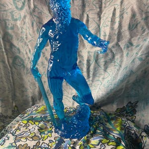 K&ouml;nnte beinhalten: Eine durchscheinende blaue Figur eines b&auml;rtigen Mannes, m&ouml;glicherweise eine mythologische Figur, die einen Stab h&auml;lt. Die Figur steht auf einer wellenartigen Basis. Die Figur ist etwa 15 cm gro&szlig;.