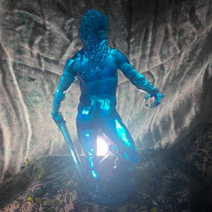 K&ouml;nnte beinhalten: Eine durchscheinende blaue Skulptur einer muskul&ouml;sen m&auml;nnlichen Figur mit langem Bart, die einen Stab h&auml;lt. Die Figur ist beleuchtet und erzeugt einen Leuchteffekt. Die Skulptur befindet sich auf einem Stoffhintergrund mit Muster.
