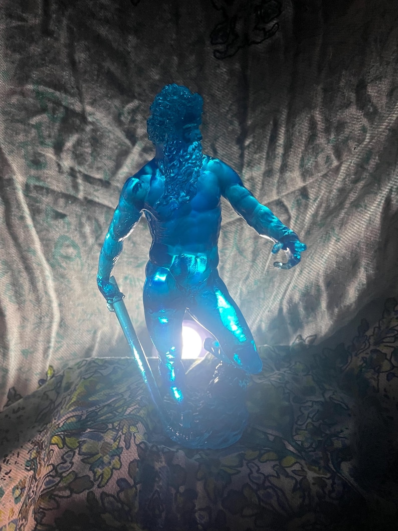 K&ouml;nnte beinhalten: Eine durchscheinende blaue Skulptur einer muskul&ouml;sen m&auml;nnlichen Figur mit langem Bart, die einen Stab h&auml;lt. Die Figur ist beleuchtet und erzeugt einen Leuchteffekt. Die Skulptur befindet sich auf einem Stoffhintergrund mit Muster.