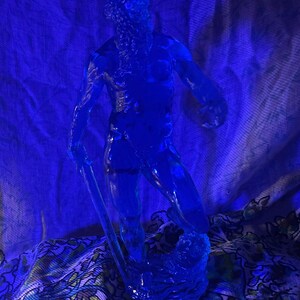K&ouml;nnte beinhalten: Eine durchscheinende blaue Skulptur eines Mannes mit Bart, der einen gebogenen Gegenstand h&auml;lt. Die Figur steht auf einem Sockel, der an Wasser erinnert. Der Hintergrund ist ein blauer Stoff mit floralem Muster.