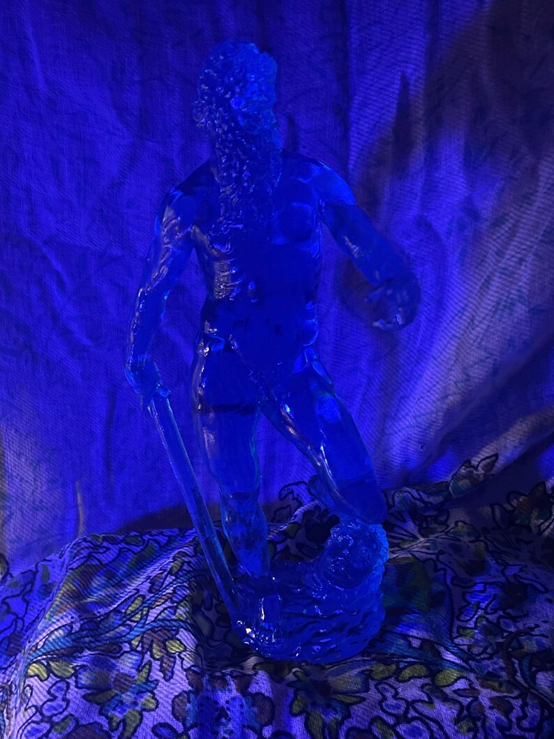 K&ouml;nnte beinhalten: Eine durchscheinende blaue Skulptur eines Mannes mit Bart, der einen gebogenen Gegenstand h&auml;lt. Die Figur steht auf einem Sockel, der an Wasser erinnert. Der Hintergrund ist ein blauer Stoff mit floralem Muster.