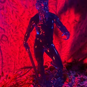 K&ouml;nnte beinhalten: Eine dunkelblaue, gl&auml;nzende Figur einer Person, die einen Stab h&auml;lt, vor rotem Hintergrund. Die Figur hat eine strukturierte Oberfl&auml;che und wird durch rote und blaue Beleuchtung beleuchtet, was einen dramatischen Effekt erzeugt. Die Figur steht auf einem gemusterten Stoff.