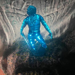 K&ouml;nnte beinhalten: Eine durchscheinende blaue Figur eines muskul&ouml;sen Mannes mit Bart, der einen Stab h&auml;lt. Die Figur ist in einer dynamischen Haltung dargestellt, wobei das Licht durch sie hindurchscheint. Die Figur ist ein dekorativer Gegenstand.
