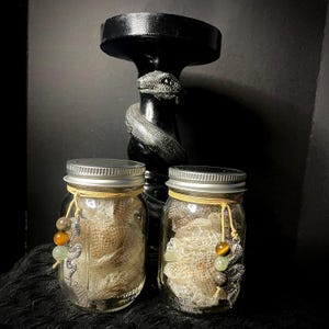 Puede incluir: Dos tarros de cristal transparente llenos de un material beige texturizado, cada uno adornado con un dije de serpiente plateada y cuentas. Un candelabro negro con un diseño de serpiente está detrás de los tarros. Los tarros tienen tapas plateadas y están atados con hilo amarillo.
