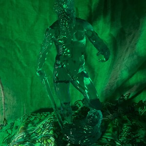 K&ouml;nnte beinhalten: Eine durchscheinende gr&uuml;ne Skulptur einer muskul&ouml;sen Figur, die eine Klinge h&auml;lt, m&ouml;glicherweise ein Krieger oder eine mythologische Figur. Die Figur ist dynamisch posiert, mit detailliertem Gesicht und K&ouml;rper. Die Skulptur steht vor einem gr&uuml;nen Stoffhintergrund mit floralem Muster.