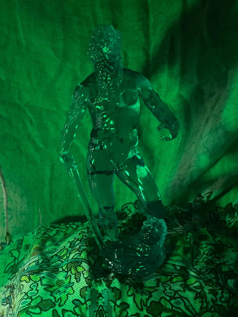 K&ouml;nnte beinhalten: Eine durchscheinende gr&uuml;ne Skulptur einer muskul&ouml;sen Figur, die eine Klinge h&auml;lt, m&ouml;glicherweise ein Krieger oder eine mythologische Figur. Die Figur ist dynamisch posiert, mit detailliertem Gesicht und K&ouml;rper. Die Skulptur steht vor einem gr&uuml;nen Stoffhintergrund mit floralem Muster.