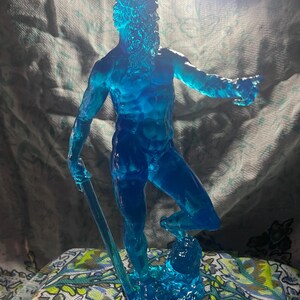 K&ouml;nnte beinhalten: Eine durchscheinende blaue Skulptur eines Mannes mit Bart, der einen Stab h&auml;lt. Die Figur ist mit einem Bein nach vorne und einem ausgestreckten Arm posiert und scheint zu zeigen. Die Skulptur steht auf einem Sockel, der Wasser &auml;hnelt.