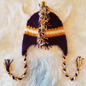 May include: Purple and gold crocheted hat with white accents and horns. The hat has a long tassel with the same colors.