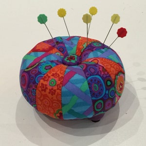 May include: A colorful, round pincushion with a patchwork design in shades of purple, orange, blue, and pink. Several pins with flower-shaped heads in yellow, green, and red are inserted into the cushion. The pincushion sits on a white surface.