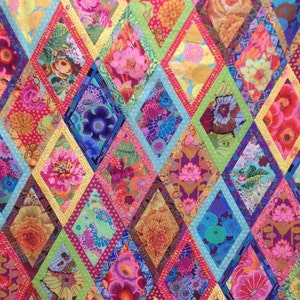 Op de afbeelding: Een levendige, kleurrijke quilt met een ruitpatroon. Elke ruit is gevuld met bloemenstof in tinten roze, blauw, geel, groen en oranje. De quilt is een patchwork ontwerp, met een verscheidenheid aan bloemenprints.