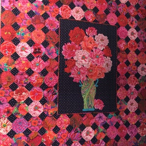 May include: A quilt with a black and white diamond pattern. The quilt is made up of many different floral fabrics in shades of pink, red, and orange. A large bouquet of pink flowers in a green vase is centered on the quilt.