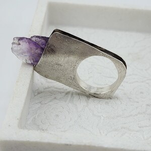 May include: A silver ring featuring a raw, uncut amethyst crystal. The ring has a rectangular face with a circular opening for the finger. The amethyst is a deep purple color and adds a natural, organic element to the design.