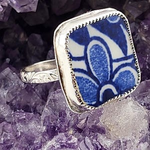 Könnte beinhalten: Ein silberner Ring mit einem quadratischen, blau-weißen Blumenmuster. Der Ring ist vor einem Hintergrund aus violetten Amethystkristallen platziert. Das Band des Rings hat ein verziertes, graviertes Muster.