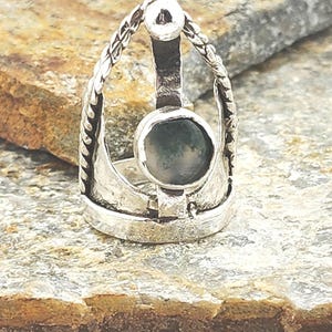 Può includere: Un anello d'argento con una pietra centrale di forma ovale. L'anello presenta un design decorativo con una fascia strutturata e una piccola sfera sulla sommità. La pietra è di colore verde tenue.