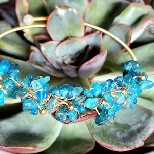 May include: A gold bangle bracelet with blue gemstone chips wrapped in gold wire. The bracelet is on a green succulent plant.