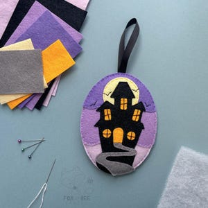 May include: A purple felt ornament featuring a black silhouette of a haunted house with orange windows. The ornament has a black ribbon for hanging and is surrounded by felt scraps in various colours.
