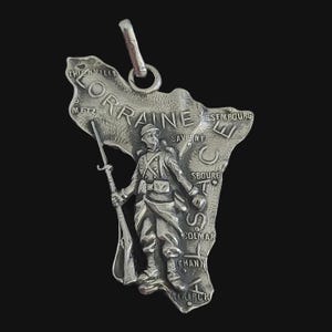 May include: A silver-toned metal pendant shaped like a map of the Alsace and Lorraine regions. The surface features a relief of a soldier in uniform holding a rifle, with engraved city names including Metz, Saverne, Strasbourg, and Colmar.