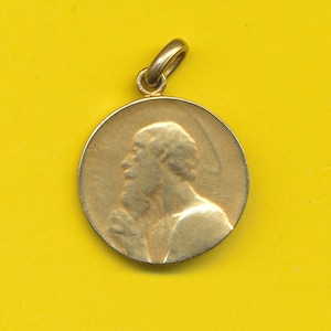 May include: A gold-colored pendant featuring a profile of a man with a beard, wearing a robe. The pendant is round and has a loop at the top for hanging.