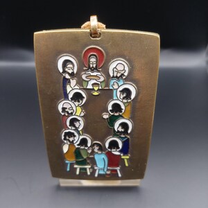May include: A rectangular gold-coloured pendant featuring a colourful enamel depiction of the Last Supper. The religious scene includes figures with halos seated around a table, with a chalice in the centre. A gold-coloured cord is attached at the top.