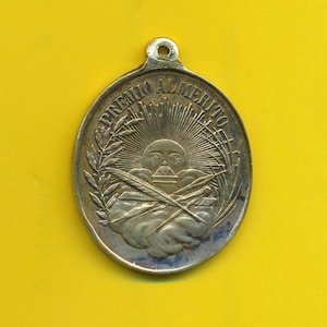 Könnte beinhalten: Eine ovale Silbermedaille mit der Aufschrift "Premio Almerito" am Rand. Die Mitte der Medaille zeigt eine Sonne mit einem Gesicht, zwei gekreuzte Federn und einen Kranz aus Blättern.