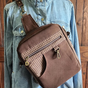 May include: A brown crossbody sling bag featuring a dark brown and tan gingham check pattern on the front pocket and top trim. The bag includes a zippered main compartment, a front zipper pocket, brass-toned hardware, and an adjustable strap.