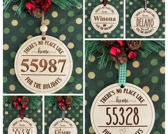 May include: Wooden Christmas ornaments with engraved text. The ornaments feature the phrase "There's no place like home" and a town name or postcode. Each ornament is adorned with a decorative ribbon and festive embellishments.