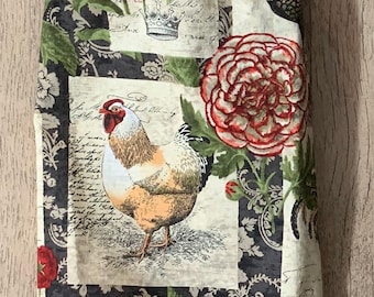 Puede incluir: Un soporte de tela para paños de cocina con lazo y cierre de botón. El soporte presenta un patrón repetido de peonías rojas, gallos y escritura sobre un fondo crema y negro. Un accesorio de cocina decorativo.