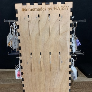 May include: Wooden display stand with the text "Homemades by HASSY" engraved on the front. The stand has multiple hooks for hanging small items, such as keychains or earrings.