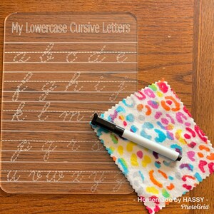 May include: A clear plastic dry-erase board with lowercase cursive letters for tracing. The board has lines for writing and a white dry-erase marker is on a colourful fabric next to the board.