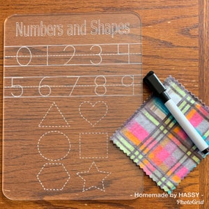 May include: A clear plastic dry-erase board with dotted lines for tracing numbers 0-9 and shapes including a triangle, heart, circle, square, hexagon, and star. The board is on a wooden surface with a dry-erase marker and a colourful fabric next to it. The text "Numbers and Shapes" is at the top of the board. The text "Homemade by HASSY" is at the bottom of the board.