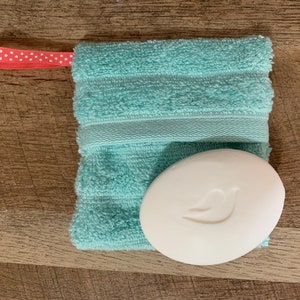 May include: A white bar of soap with a bird logo is inside a light blue washcloth with a red and white polka dot ribbon.