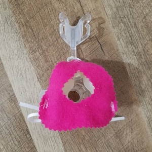 Può includere: Una maschera facciale in tessuto rosa con un pezzo nasale in plastica trasparente e lacci elastici bianchi.