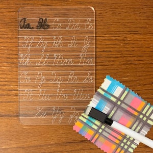 May include: A clear plastic dry-erase board with cursive alphabet letters printed on it. The board is on a wooden surface. A white dry-erase marker and a colourful fabric are also in the image.