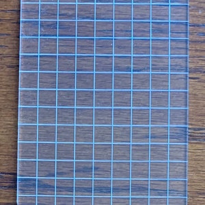 May include: A clear acrylic grid sheet with a white grid pattern. The grid is evenly spaced and has a total of 100 squares.